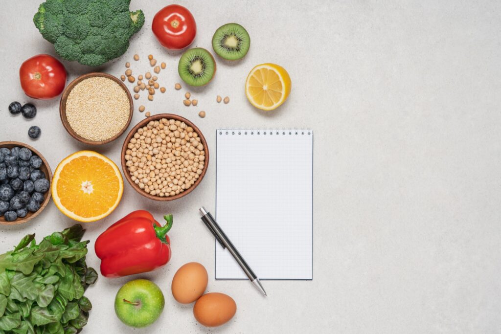 Plan de alimentación para la diabetes tipo 2: Variedad de alimentos saludables junto a un bloc de notas para planificar las comidas. La planificación de comidas ayuda a controlar la glucosa en sangre en la diabetes tipo 2.
