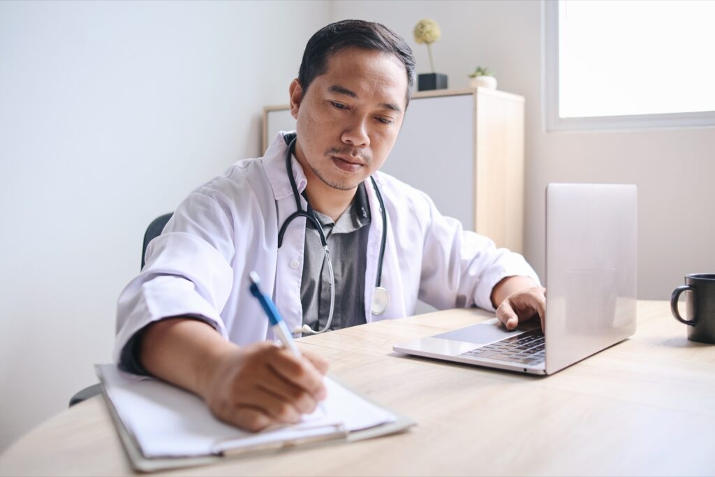 Un médico revisa el historial médico de un paciente con AME en su ordenador portátil y toma notas. Representando la importancia de la investigación y el desarrollo de tratamientos para la AME.