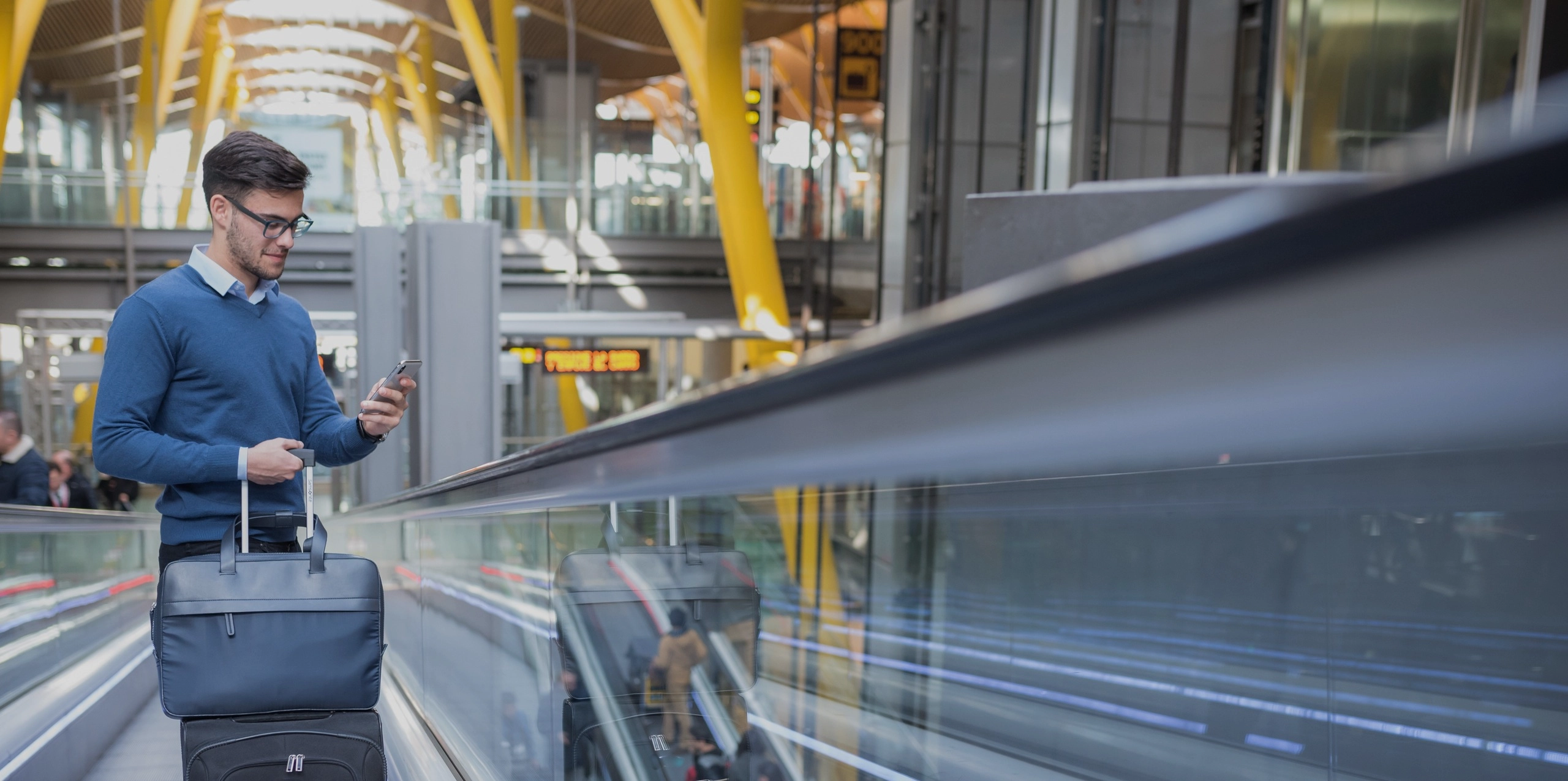 Joven en la escalera mecanica en el aeropuerto usando su telefono movil con su equipaje sonriendo 1 2560x1276 1