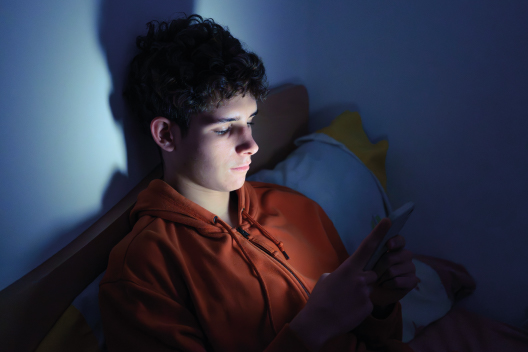 Chico joven en una habitación a oscuras iluminado únicamente por la luz de la pantalla de su teléfono móvil.
