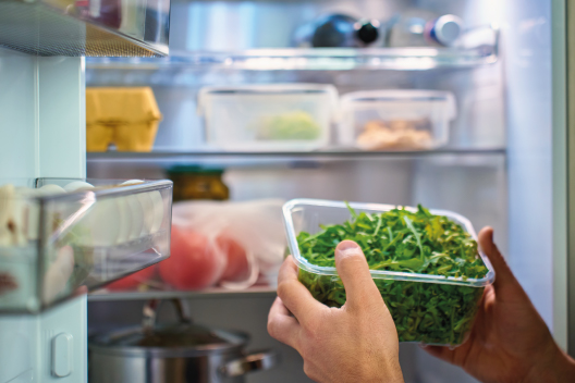 Personas guardando un recipiente de plástico con vegetales verdes dentro de una nevera abierta llena de alimentos.