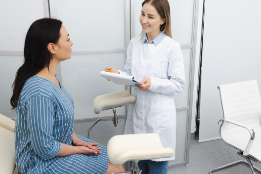 Médica especialista con bata blanca y carpeta informando a una paciente sentada en la camilla durante una revisión ginecológica.